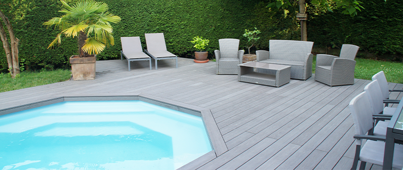 Große Poolterrasse mit grauem WPC-Belag, modernen Lounge-Möbeln und Liegestühlen, umgeben von grüner Hecke und Palmen.