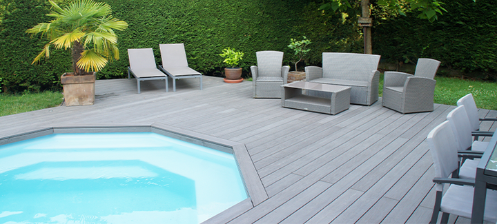Große Poolterrasse mit grauem WPC-Belag, modernen Lounge-Möbeln und Liegestühlen, umgeben von grüner Hecke und Palmen.