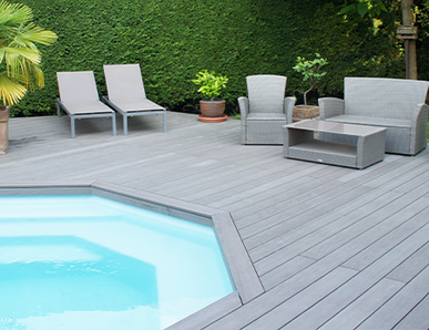 Große Poolterrasse mit grauem WPC-Belag, modernen Lounge-Möbeln und Liegestühlen, umgeben von grüner Hecke und Palmen.